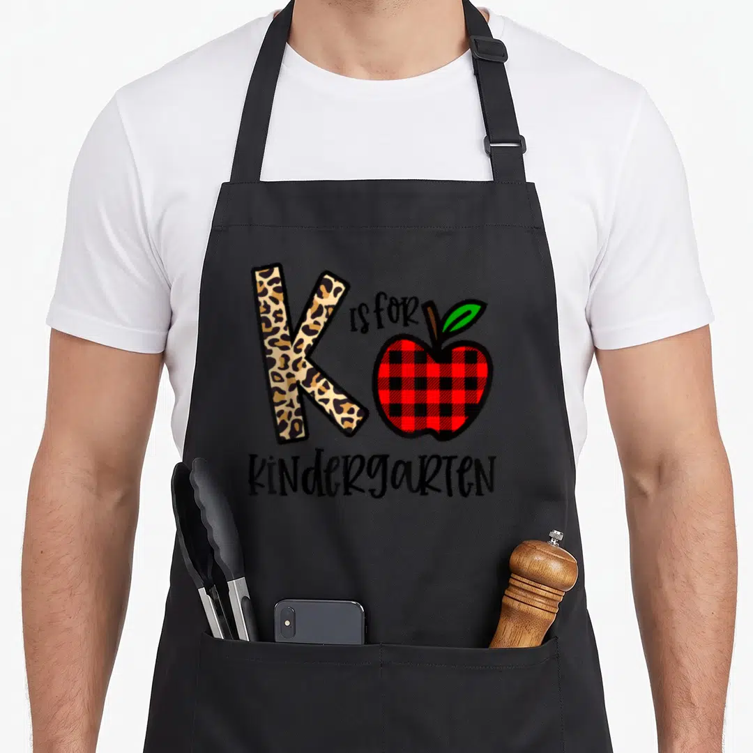 Leopard kindergarten teacher apron - Leopard print letter K next to red plaid apple and black KINDERGARTEN text — shown on a black cotton canvas apron against a white background