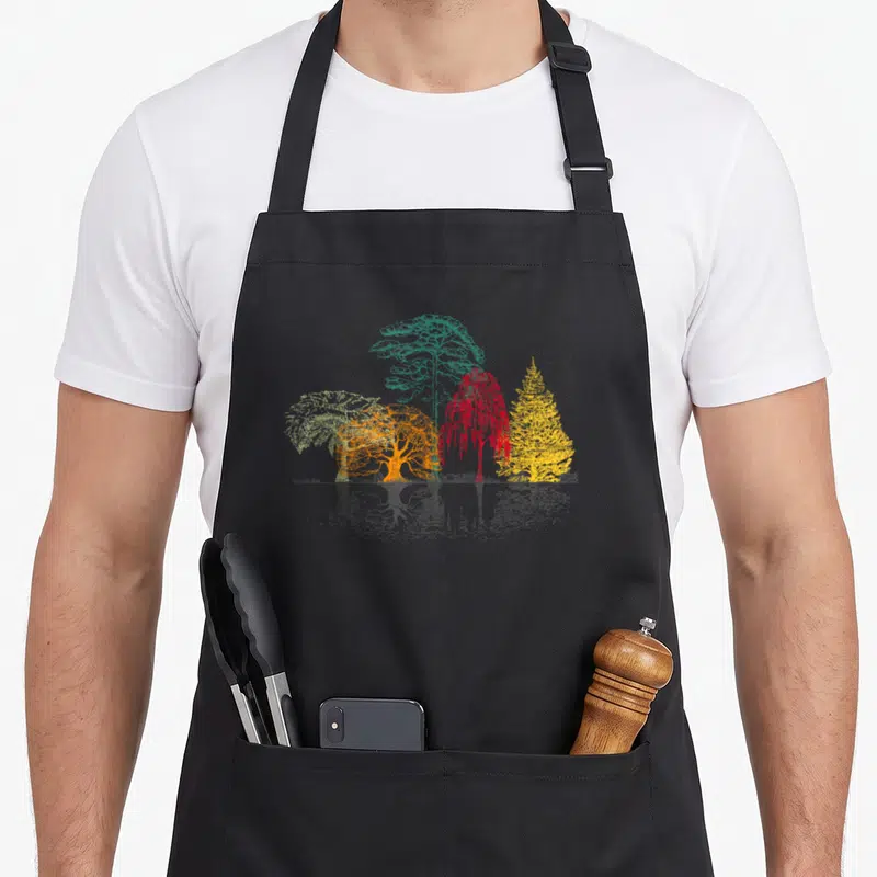 Nature outdoor apron - Four colorful trees in green orange red yellow with grey reflection below — shown on a black cotton canvas apron against a white background