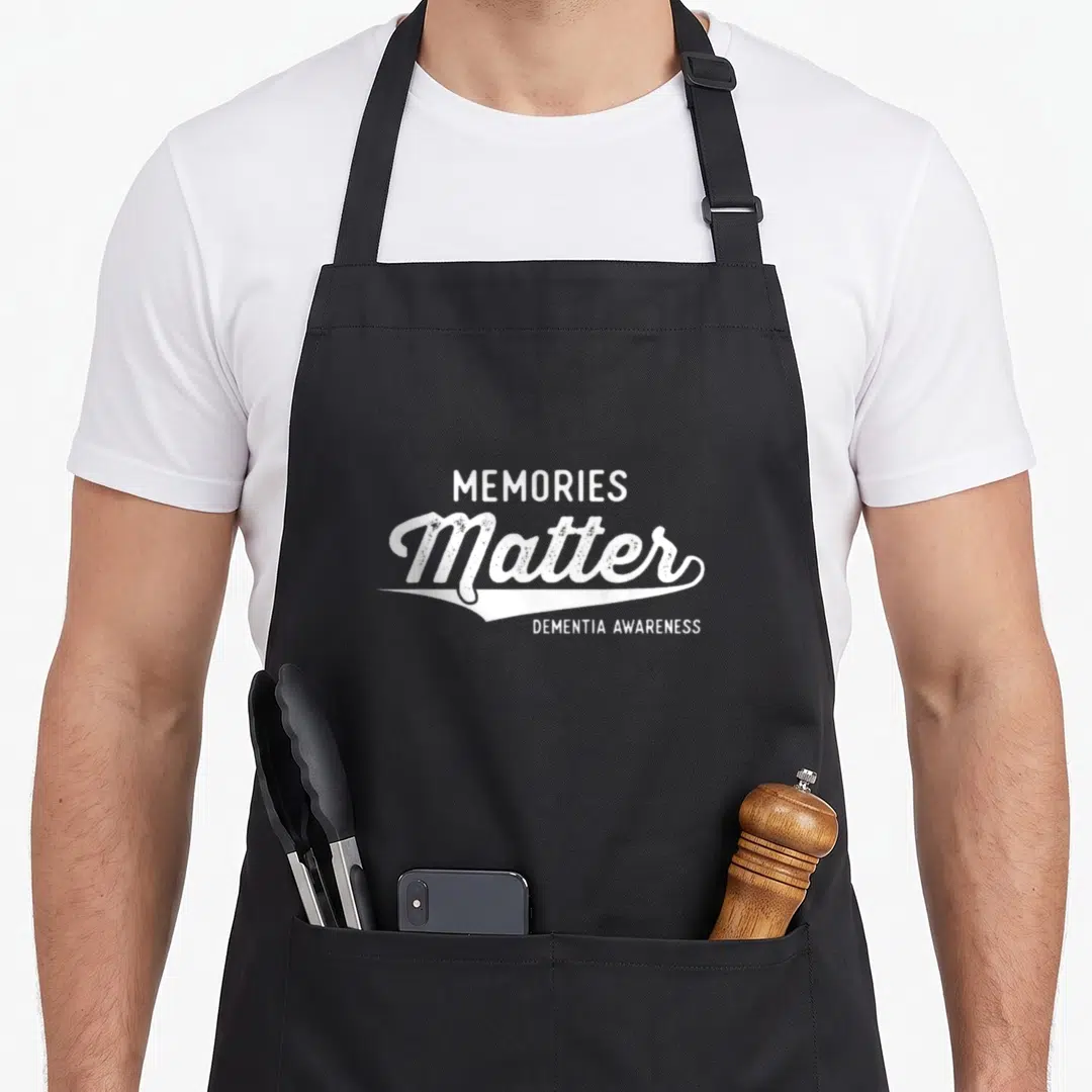 Awareness memory apron - White stacked text MEMORIES Matter DEMENTIA AWARENESS with decorative swoosh on black fabric — shown on a black cotton canvas apron against a white background