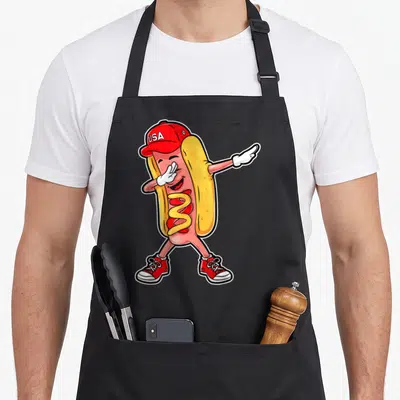 Merica hot dog apron - Cartoon hot dog character dabbing wearing USA cap and red sneakers with mustard drizzle — shown on a black cotton canvas apron against a white background