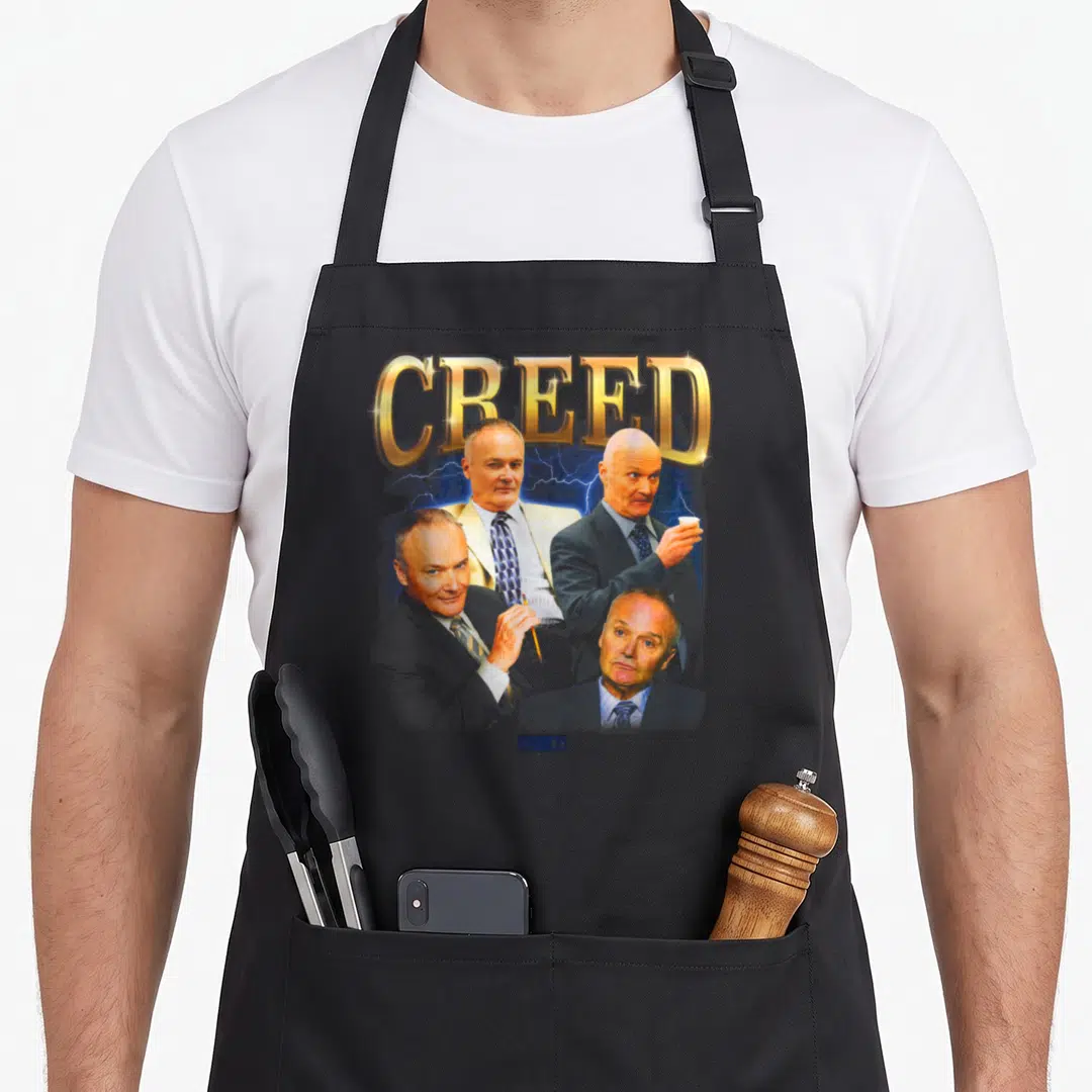 Creed nbc apron - Gold CREED text above four portraits of a man against blue lightning on grey background — shown on a black cotton canvas apron against a white background