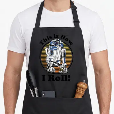Funny roll apron - R2-D2 in black circle with orange stripes and This Is How I Roll text — shown on a black cotton canvas apron against a white background
