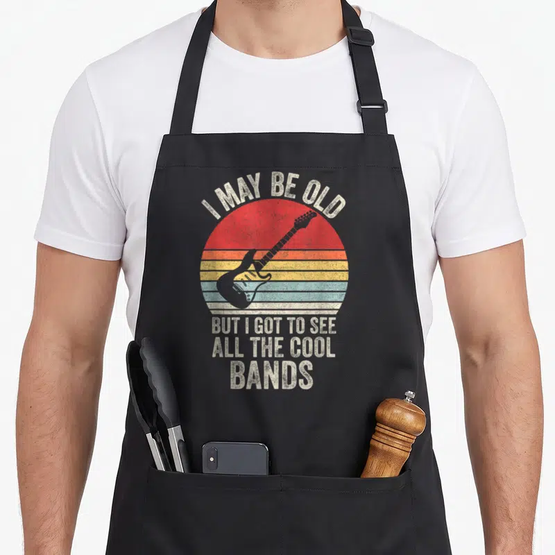 See band may apron - Electric guitar over red orange yellow teal striped sun with I MAY BE OLD text — shown on a black cotton canvas apron against a white background