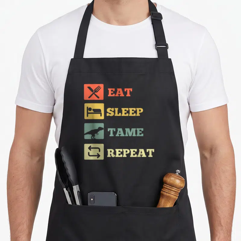 Sleep dinosaur apron - Four rows of text with colored squares showing cutlery, bed, dinosaur, and arrows — shown on a black cotton canvas apron against a white background