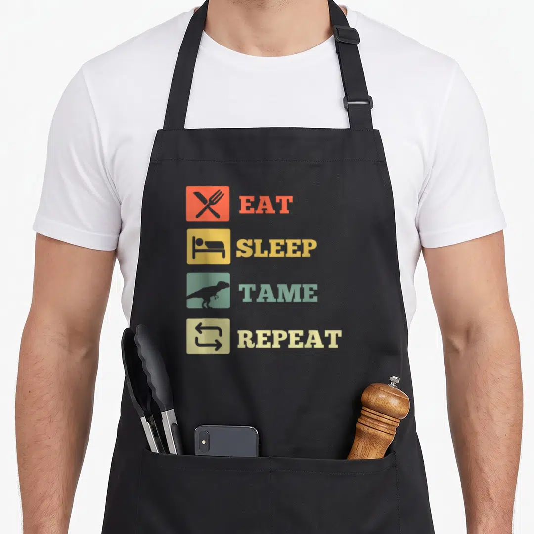 Sleep dinosaur apron - Four rows of text with colored squares showing cutlery, bed, dinosaur, and arrows — shown on a black cotton canvas apron against a white background