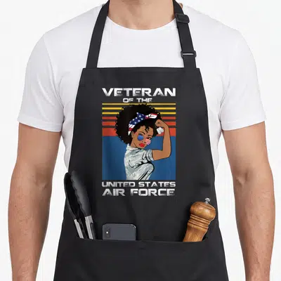 State female veteran apron - Retro woman in USAF camo holding dog tags with flag sunglasses against striped sunset — shown on a black cotton canvas apron against a white background