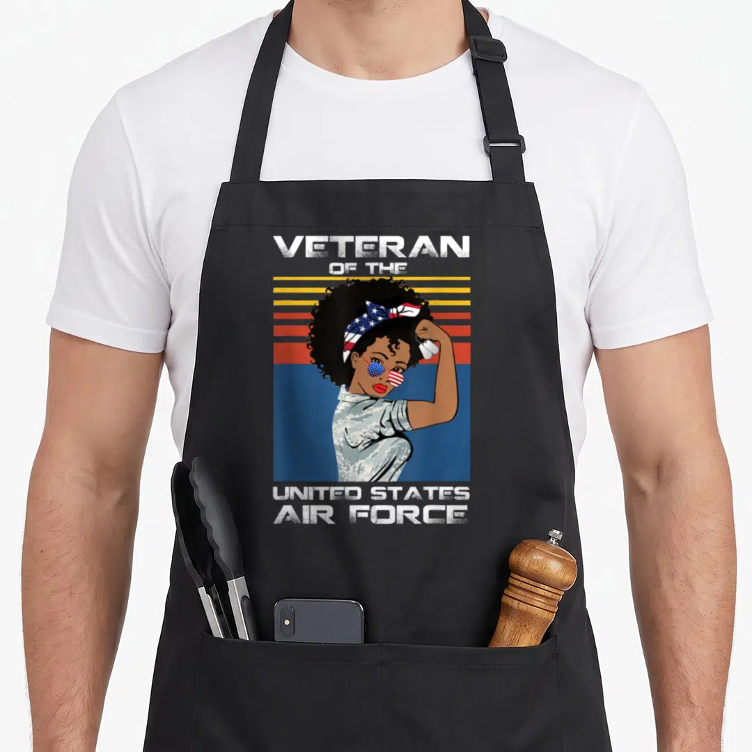 State female veteran apron - Retro woman in USAF camo holding dog tags with flag sunglasses against striped sunset — shown on a black cotton canvas apron against a white background