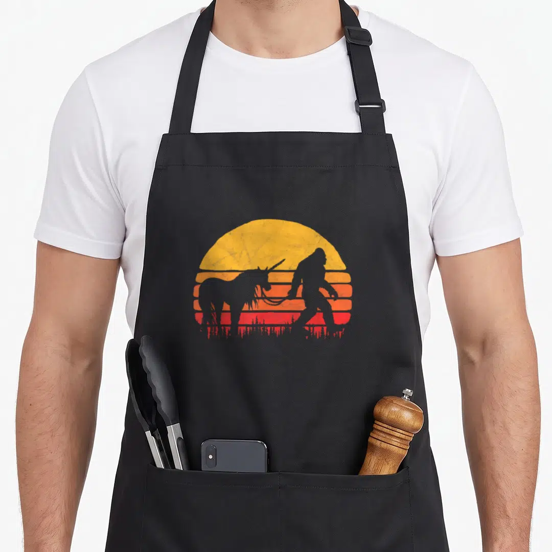 Unicorn bigfoot apron - Bigfoot silhouette holding unicorn leash against yellow orange retro striped sun — shown on a black cotton canvas apron against a white background