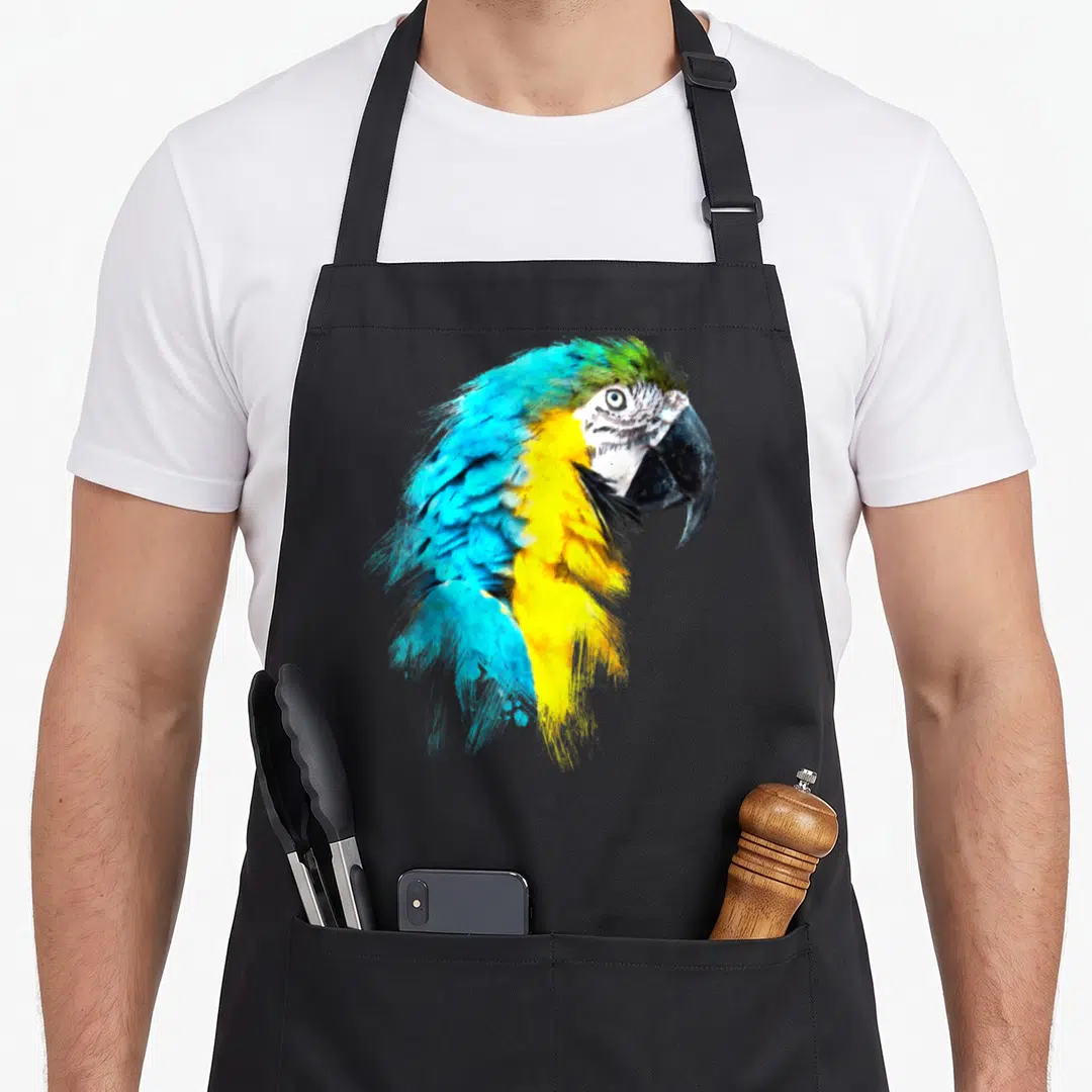 Colourful bird apron - Watercolor macaw head illustration in blue, yellow, and green feathers — shown on a black cotton canvas apron against a white background