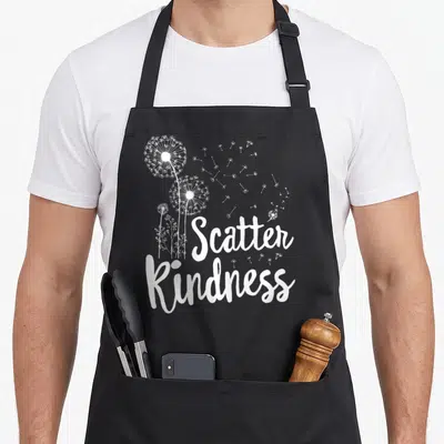 Unity kindness teacher apron - Three white dandelions with scattering seeds and script text reading Scatter Kindness — shown on a black cotton canvas apron against a white background