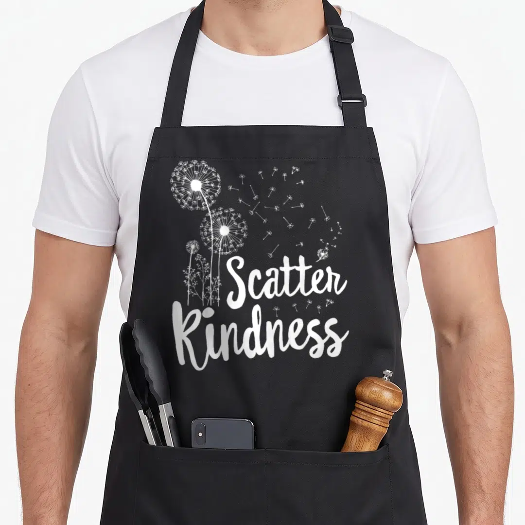 Unity kindness teacher apron - Three white dandelions with scattering seeds and script text reading Scatter Kindness — shown on a black cotton canvas apron against a white background