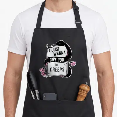 Give goth apron - Black snake coiled grey tombstone pink flowers I JUST WANNA WANNA GIVE YOU THE CREEPS text cartoon — shown on a black cotton canvas apron against a white background