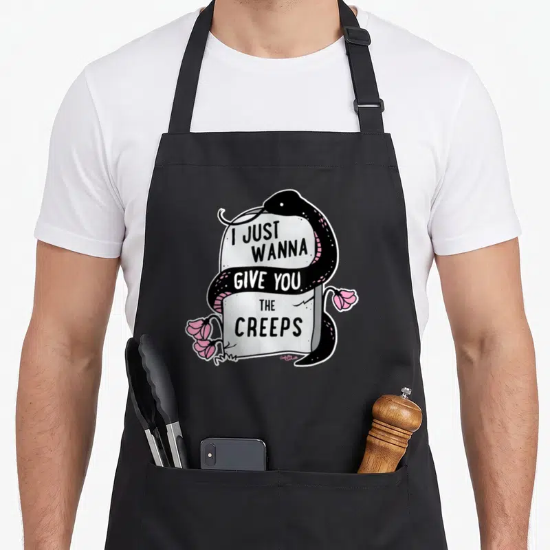 Give goth apron - Black snake coiled grey tombstone pink flowers I JUST WANNA WANNA GIVE YOU THE CREEPS text cartoon — shown on a black cotton canvas apron against a white background