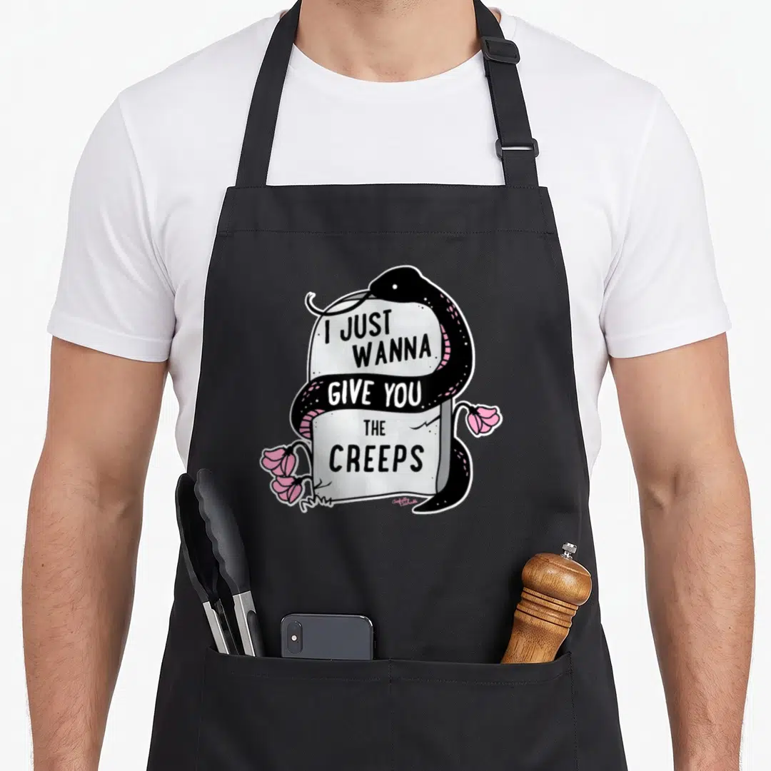 Give goth apron - Black snake coiled grey tombstone pink flowers I JUST WANNA WANNA GIVE YOU THE CREEPS text cartoon — shown on a black cotton canvas apron against a white background