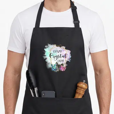 Crystal stone apron - Stacked text above purple blue pink and gold crystals in gold frame on watercolor splash — shown on a black cotton canvas apron against a white background