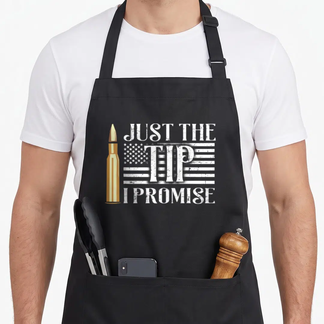 Flag gun apron - Vertical gold bullet next to distressed white text reading Just The Tip I Promise over a USA flag — shown on a black cotton canvas apron against a white background