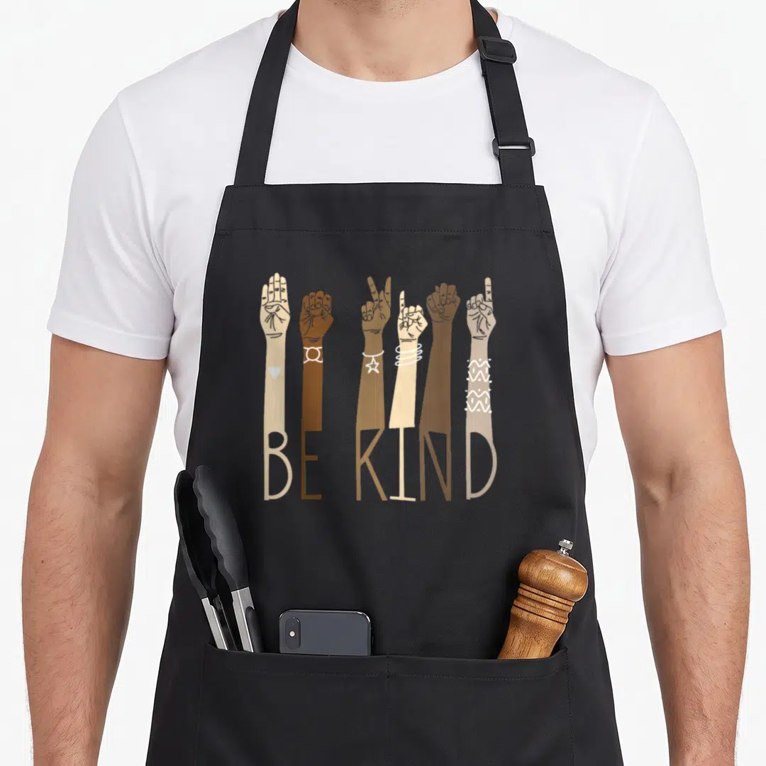 Kind asl sign for apron - Six diverse arms in beige and brown forming the words BE KIND through ASL lettering — shown on a black cotton canvas apron against a white background