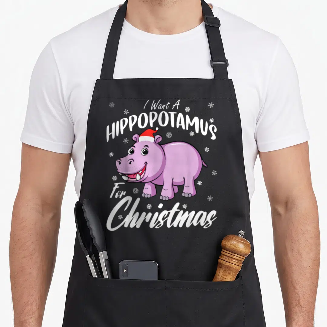 Hippo christmas apron - Purple cartoon hippo in red Santa hat with white snowflakes and arched text — shown on a black cotton canvas apron against a white background