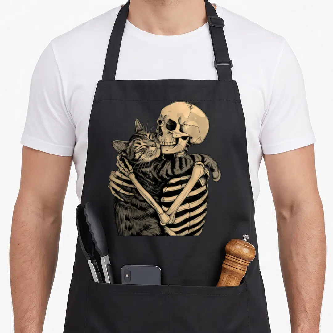Skeleton cat in an apron - Line art of a skeleton hugging a striped tabby cat in beige and black ink — shown on a black cotton canvas apron against a white background