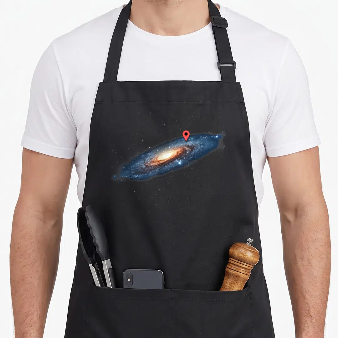 Milky pins for aprons - Spiral galaxy with red location pin and white stars on black background — shown on a black cotton canvas apron against a white background