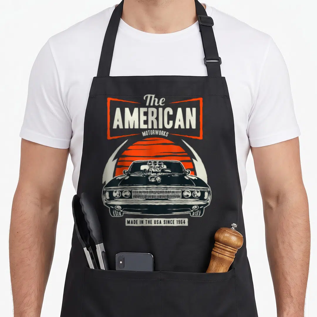 Funny car muscle apron - Black hot rod car front view with orange striped sun and retro typography reading The AMERICAN — shown on a black cotton canvas apron against a white background