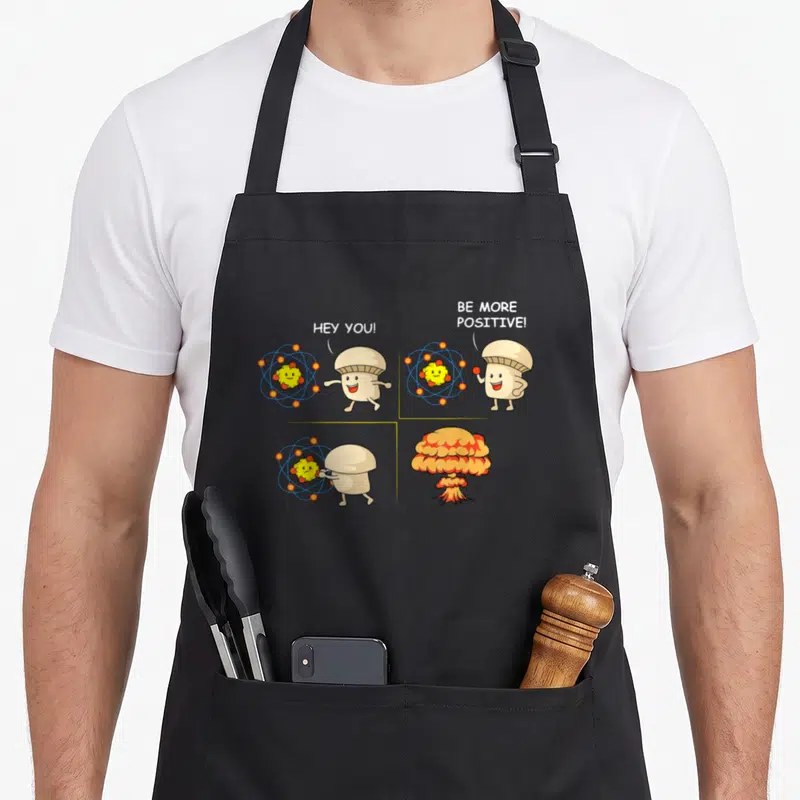 Student science apron - Mushroom character holding atom in four-panel comic ending in explosion with text HEY YOU BE — shown on a black cotton canvas apron against a white background
