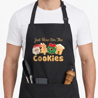 Santa women's christmas aprons - Just Here For The Cookies text with Santa, tree, wreath, and gingerbread house cookies — shown on a black cotton canvas apron against a white background