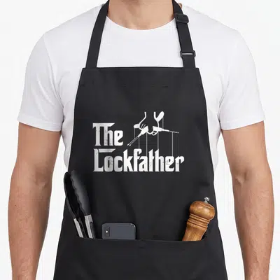 Lockfather locksmith squad apron - White distressed text The Lockfather with hand holding pick — shown on a black cotton canvas apron against a white background