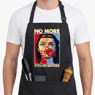 Awareness native american apron - No More Stolen Sisters text surrounding a stylized woman's portrait in red, blue — shown on a black cotton canvas apron against a white background