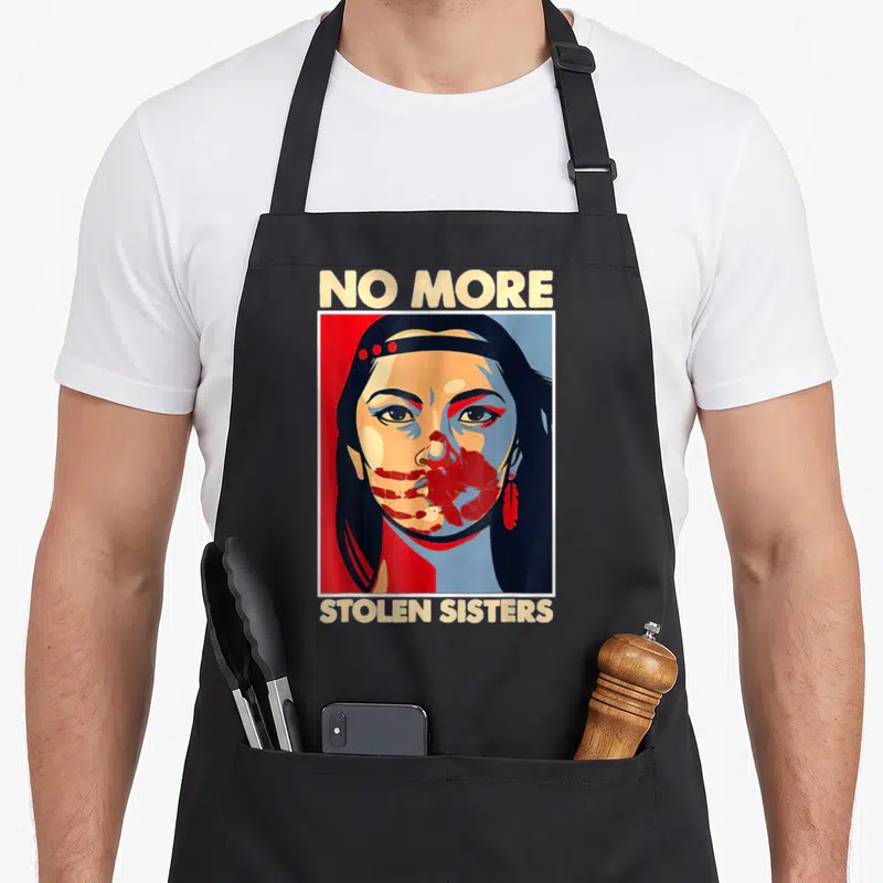 Awareness native american apron - No More Stolen Sisters text surrounding a stylized woman's portrait in red, blue — shown on a black cotton canvas apron against a white background