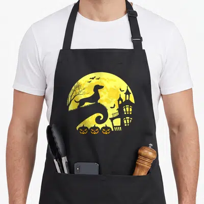 Halloween sausage apron - Grey dachshund silhouette yellow moon three orange pumpkins haunted house — shown on a black cotton canvas apron against a white background