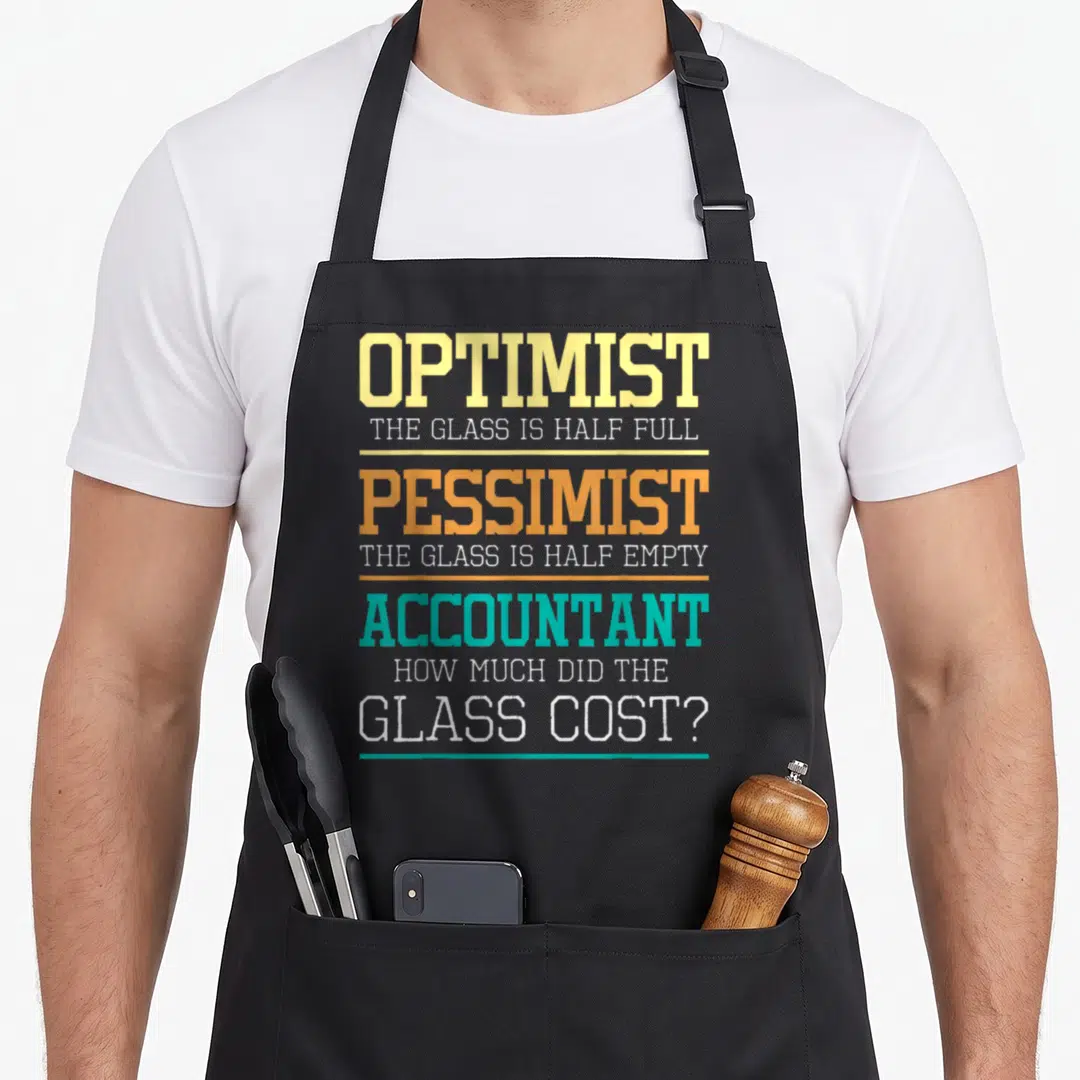 Optimist joke apron - Stacked text reading Optimist Pessimist Accountant with glass cost pun in yellow orange teal — shown on a black cotton canvas apron against a white background