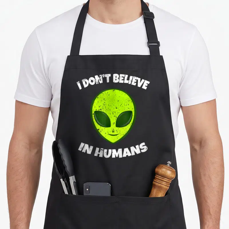 Believe funny alien apron - Lime green alien head framed by arched white text reading I Don't Believe in Humans — shown on a black cotton canvas apron against a white background