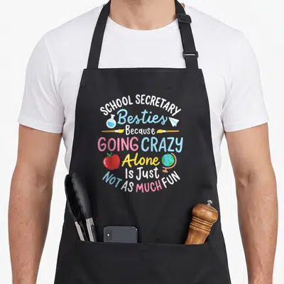 School secretary apron - Stacked colorful text reading School Secretary Besties with icons including apple, globe — shown on a black cotton canvas apron against a white background