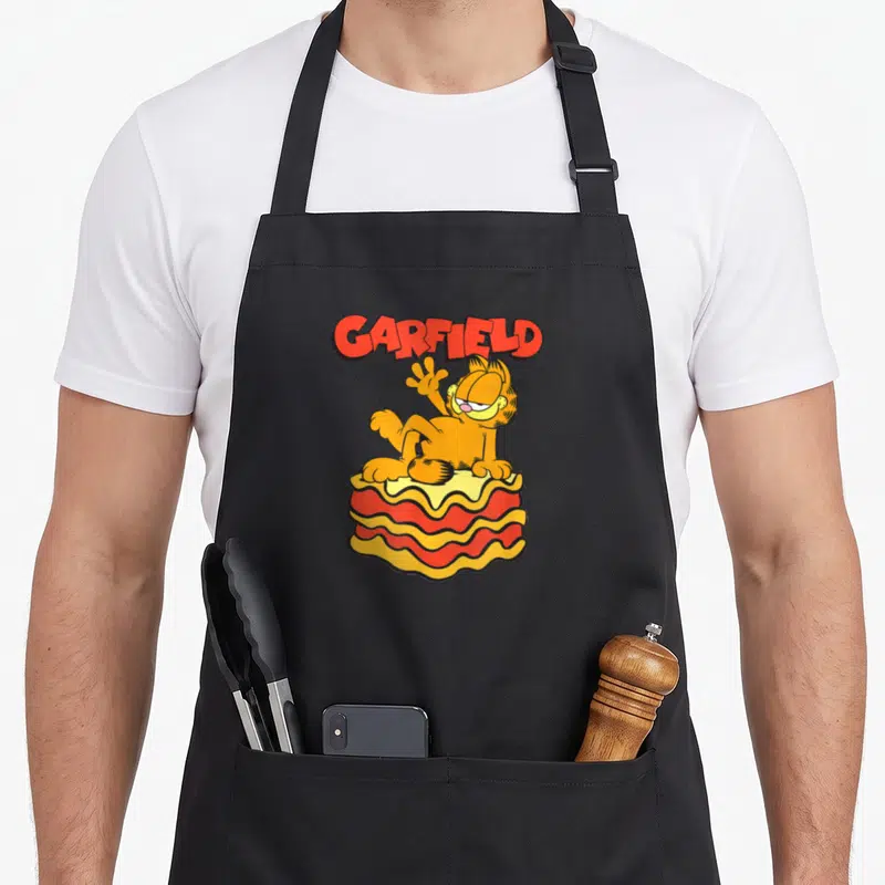 Lasagna general apron - Orange cartoon cat sitting on a wavy lasagna slice under red GARFIELD text — shown on a black cotton canvas apron against a white background