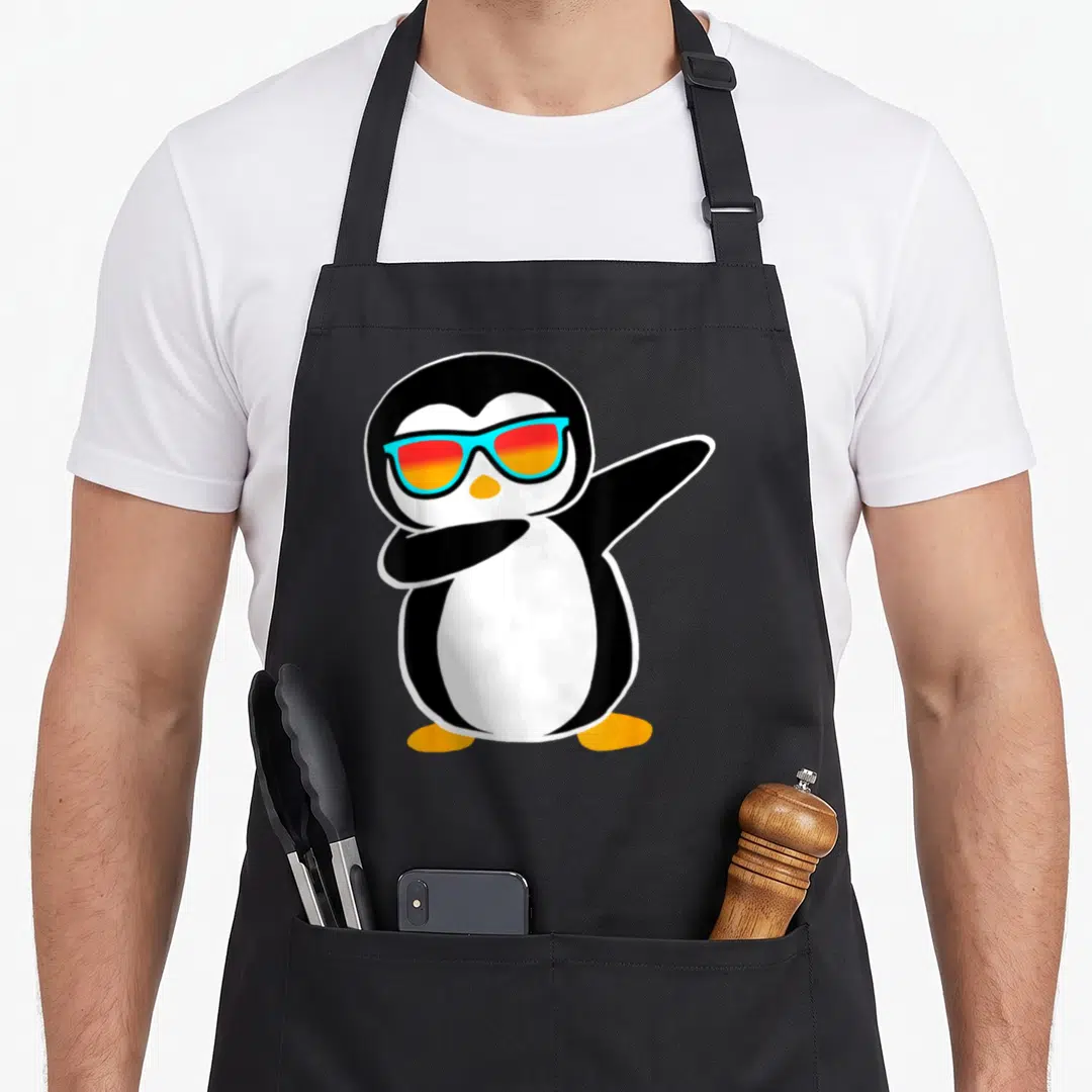 Birthday penguin aprons - Cartoon penguin dabbing with blue sunglasses and orange feet — shown on a black cotton canvas apron against a white background