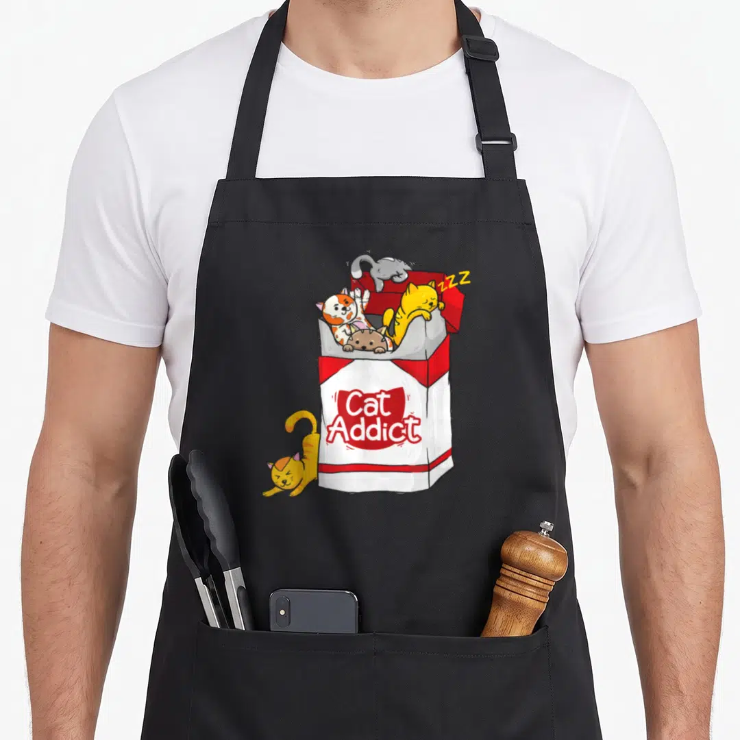 Addict cat apron - Cartoon red and white cigarette pack with four cats spilling out and one lying beside it — shown on a black cotton canvas apron against a white background
