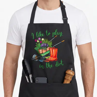 Play gardener apron - Green text arching over a wheelbarrow with potted purple and red flowers and orange boots — shown on a black cotton canvas apron against a white background