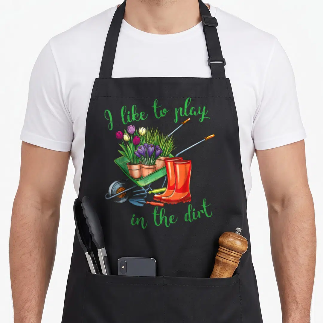 Play gardener apron - Green text arching over a wheelbarrow with potted purple and red flowers and orange boots — shown on a black cotton canvas apron against a white background