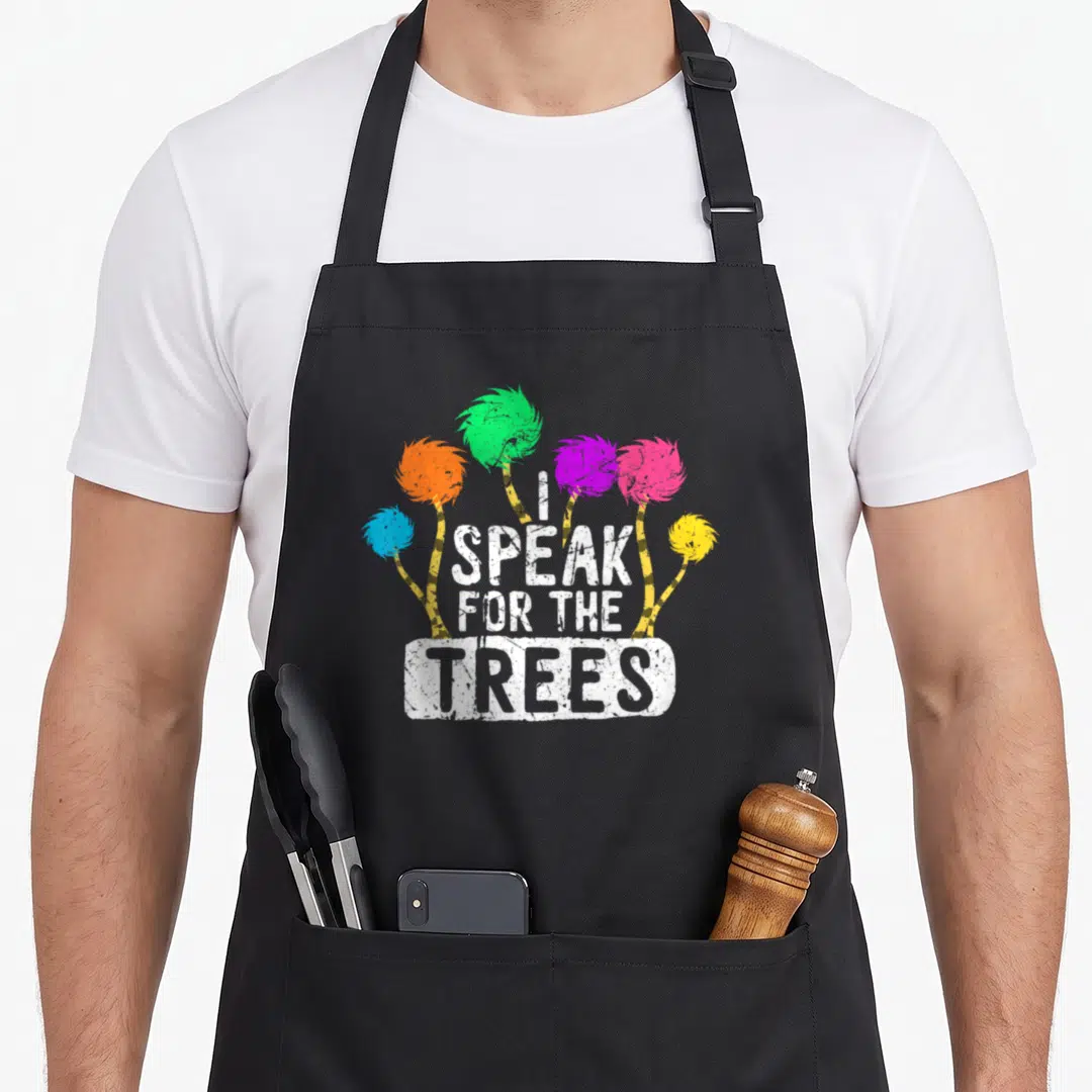 Save tree apron - Six colorful cartoon trees behind stacked white text I SPEAK FOR THE TREES — shown on a black cotton canvas apron against a white background