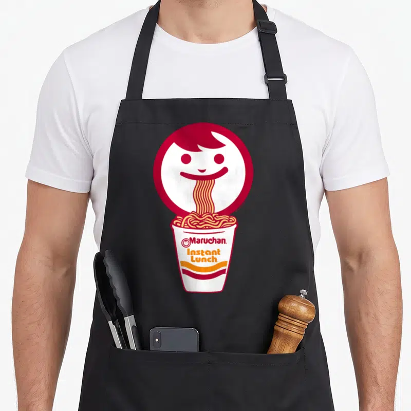 Eating face apron - Red cartoon face slurping noodles into a Maruchan Instant Lunch cup — shown on a black cotton canvas apron against a white background