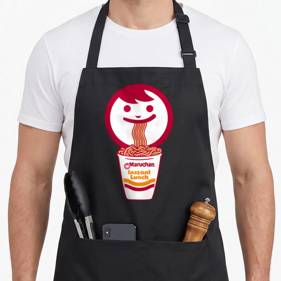 Eating face apron - Red cartoon face slurping noodles into a Maruchan Instant Lunch cup — shown on a black cotton canvas apron against a white background