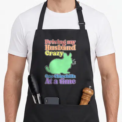 Chinchilla husband driving apron - Pastel text reading Driving my Husband Crazy One Chinchilla At a time above a green — shown on a black cotton canvas apron against a white background