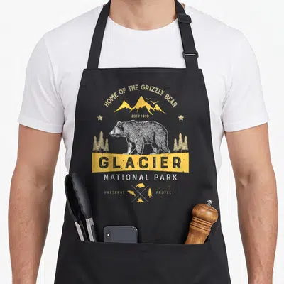 National park bear apron - Grizzly bear illustration under yellow mountains with GLACIER banner and preserve protect — shown on a black cotton canvas apron against a white background