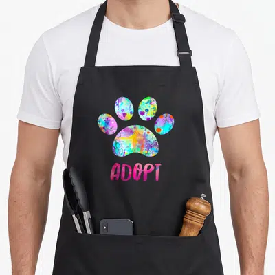 Watercolor grill rescue apron - Multicolored watercolor paw print above pink ADOPT text — shown on a black cotton canvas apron against a white background