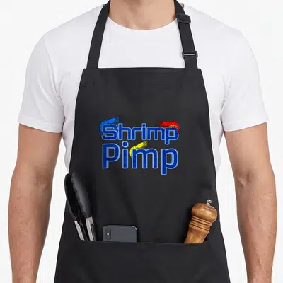 Shrimp cherry apron - Stacked Shrimp Pimp text with blue red and yellow cartoon shrimp illustrations — shown on a black cotton canvas apron against a white background