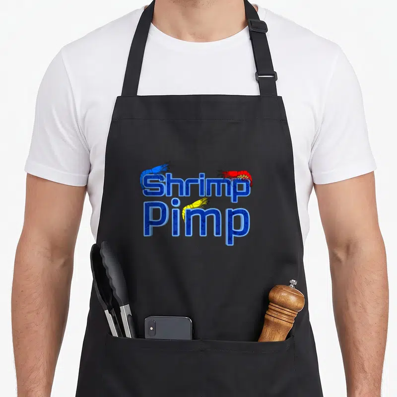 Shrimp cherry apron - Stacked Shrimp Pimp text with blue red and yellow cartoon shrimp illustrations — shown on a black cotton canvas apron against a white background