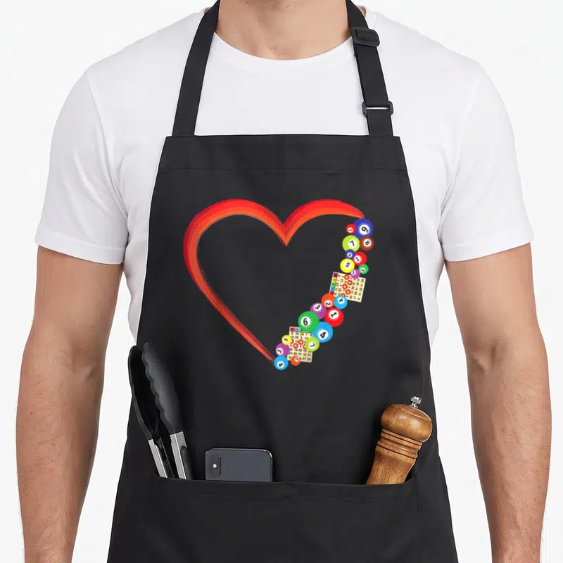 Cool player bingo apron - Red heart outline formed by colorful numbered bingo balls and cards in cartoon style — shown on a black cotton canvas apron against a white background