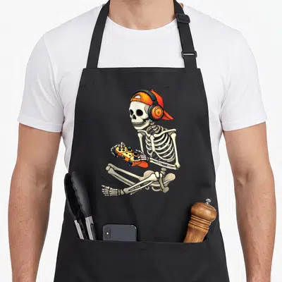 Gamer video gaming halloween apron - Cartoon skeleton sitting in profile wearing orange cap and headphones holding — shown on a black cotton canvas apron against a white background