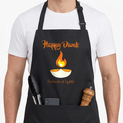 Light diwali apron - Happy Diwali text above a cartoon diya lamp with orange flame and white base — shown on a black cotton canvas apron against a white background